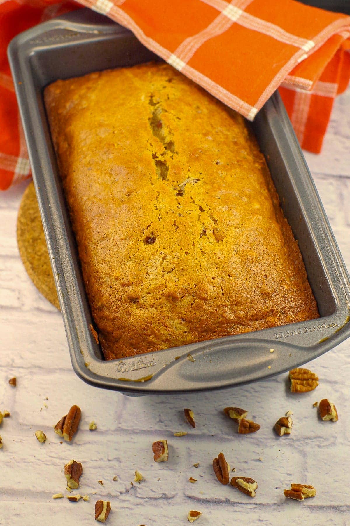 Date loaf in a pan with an orange tea towel and chopped pecans around it and