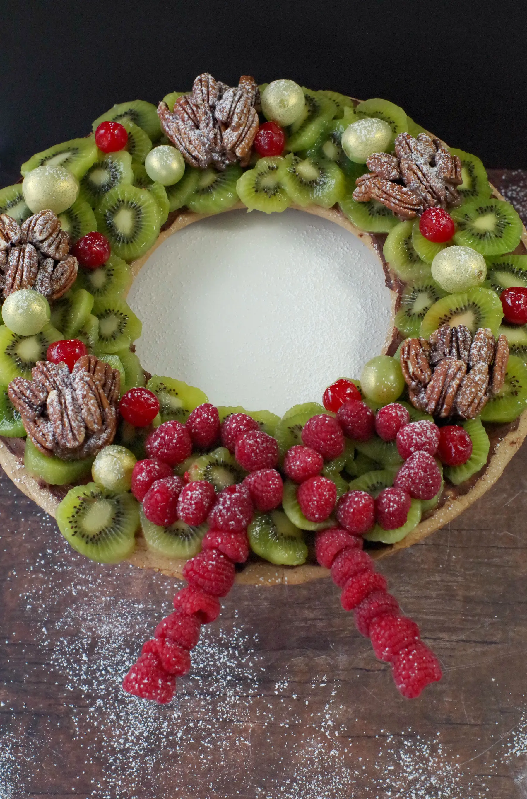 Gingerbread Edible Christmas Wreath with fruit and nuts, on brown wooden surface with powdered sugar sprinkles