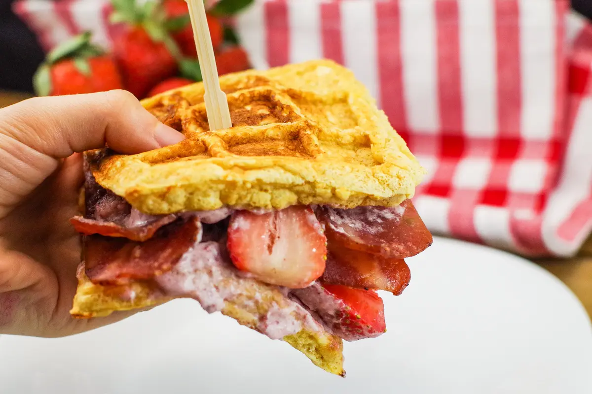 chicken and waffle sandwich being held up in a hand with strawberries in background