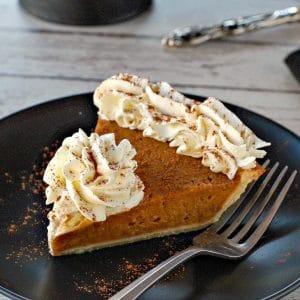 piece of pumpkin pie with whipped cream on a black plate with black platter stand in the background, with pie on platter