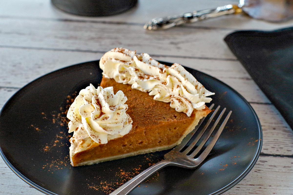 piece of pumpkin pie with whipped cream on a black plate with black platter stand in the background, with pie on platter