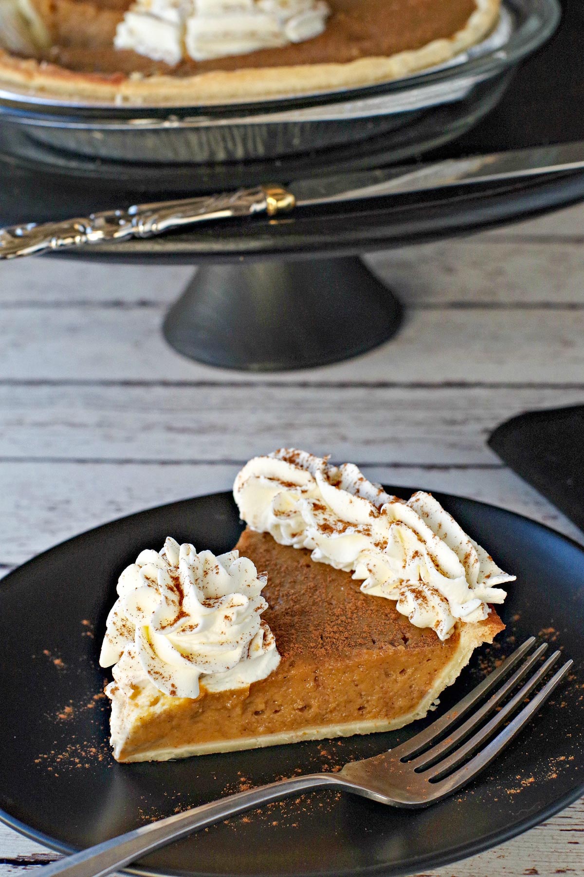 piece of pumpkin pie with whipped cream on a black plate with black platter stand in the background, with pie on platter with black platter in the background