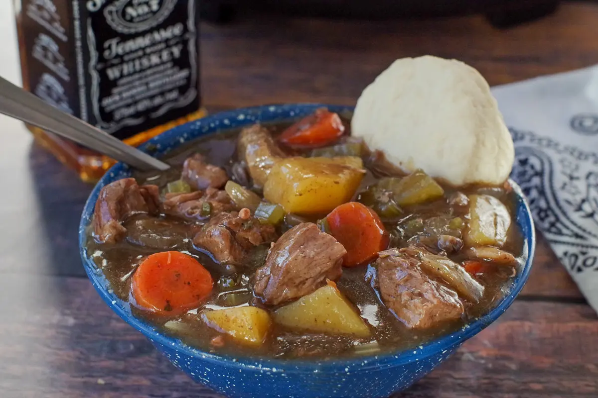 slow cooker beef stew in a blue bowl with a biscuit and slow cooker and bottle of whiskey in the background
