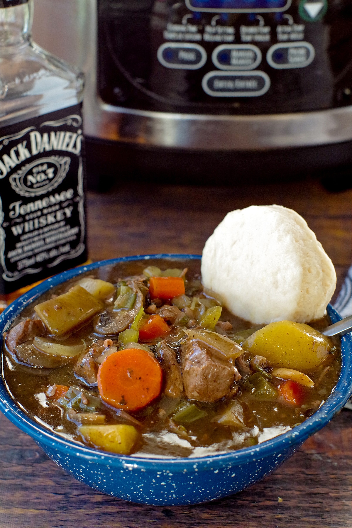 slow cooker beef stew in a blue bowl with a biscuit and slow cooker and bottle of whiskey in the background