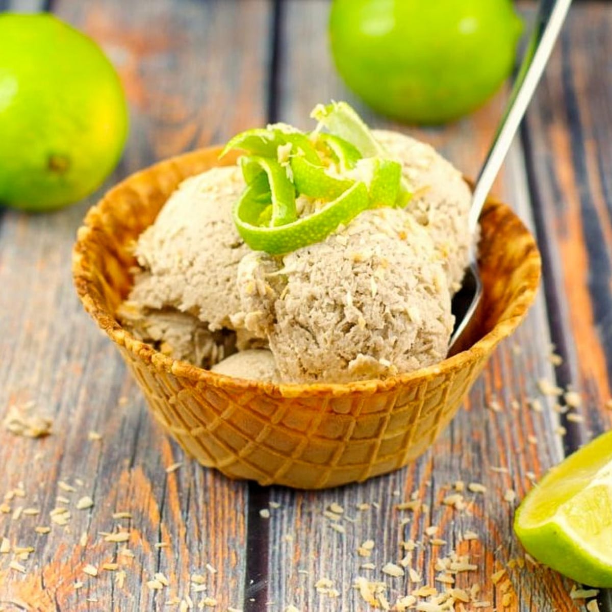 banana nice cream in a waffle cone bowl with a spoon, garnished with coconut and lime zest , with whole limes in the background