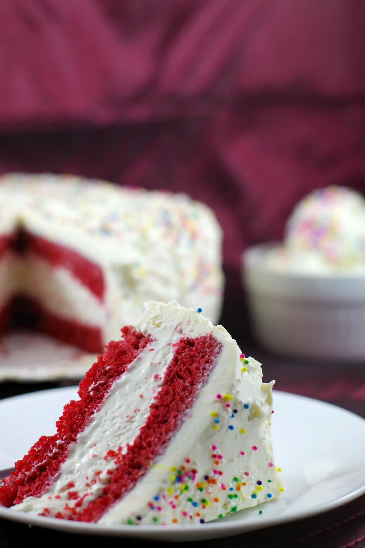 Slice of red velvet ice cream cake with whole cake in the background and a bowl of no churn ice cream