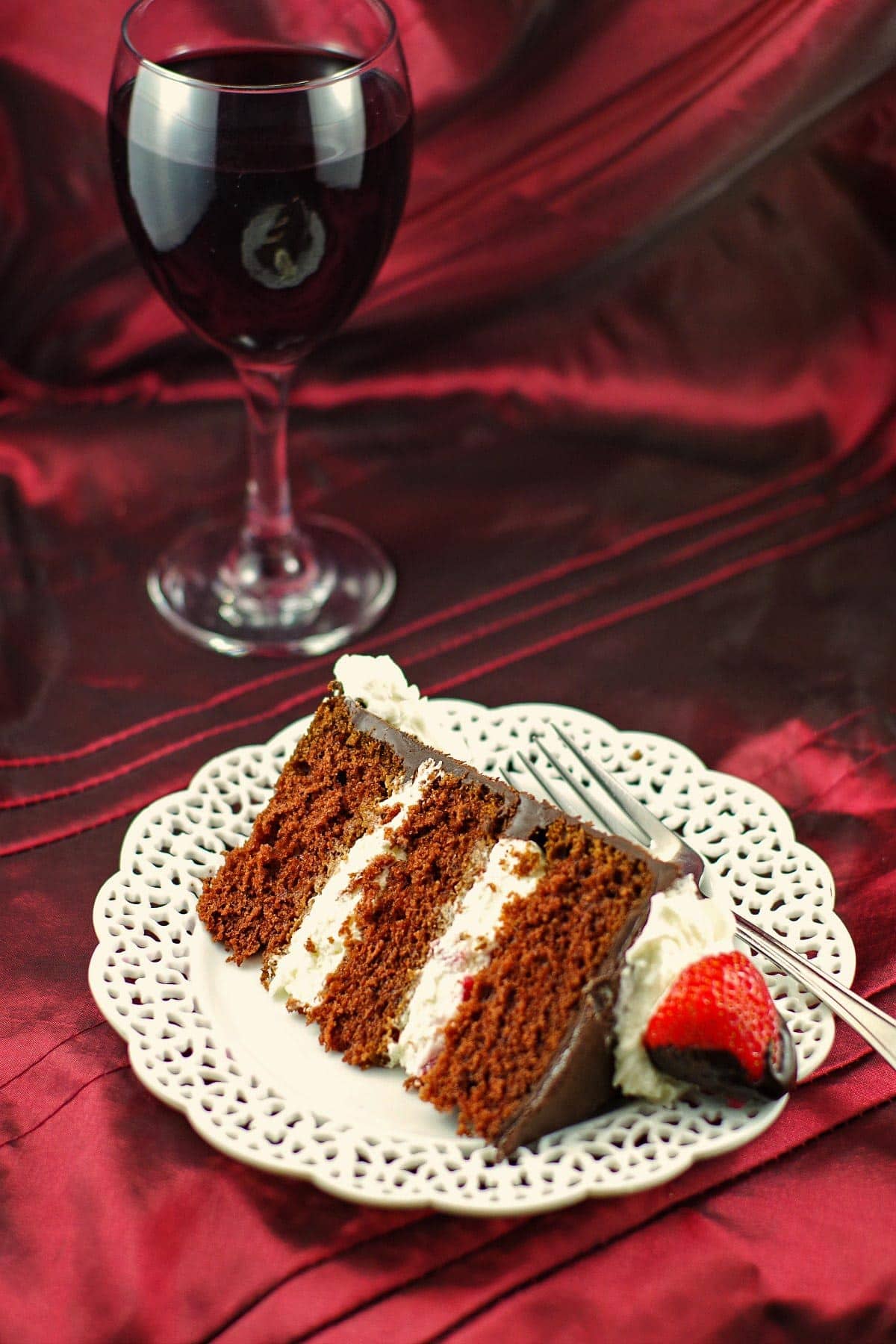 Slice of red wine chocolate strawberry cake on a white plate with glass of red wine in the background
