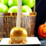 gourmet caramel apple on a white plate with basket of green apples and plastic jack o 'lantern in the background