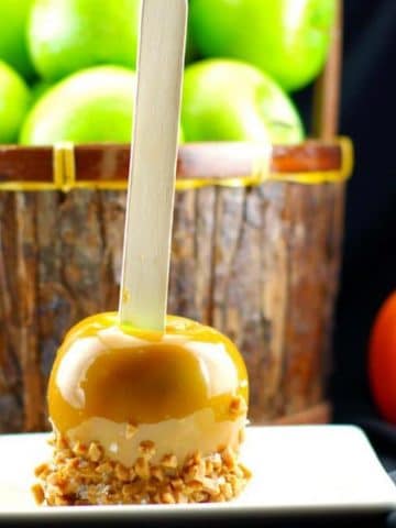 gourmet caramel apple on a white plate with basket of green apples and plastic jack o 'lantern in the background