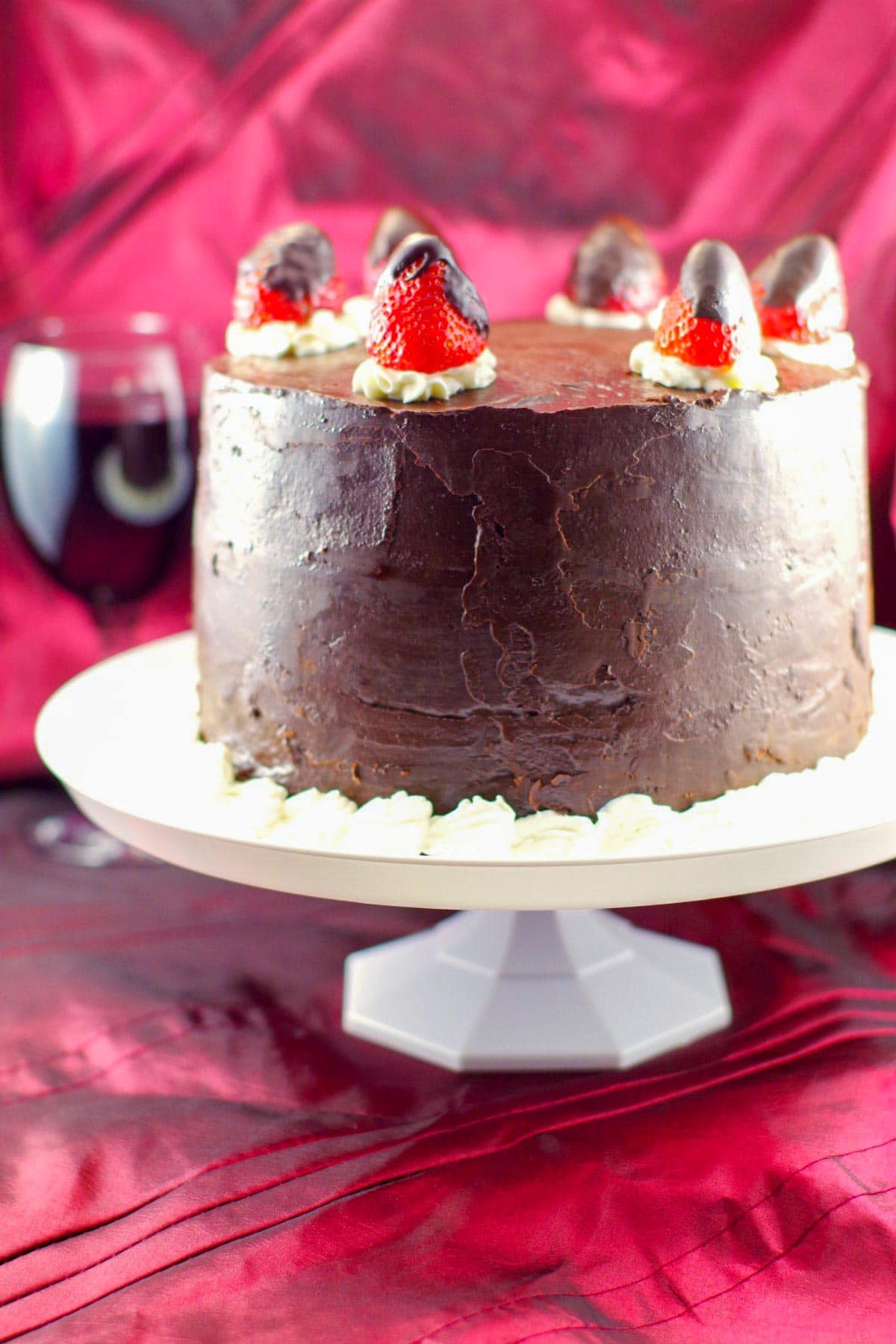 Whole red wine chocolate strawberry cake on a white platter with a glass of red wine in the background