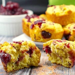 cranberry cornbread muffin split in half, with more cranberry cornbread muffins on a white plate, and a dish of cranberry sauce, in the background