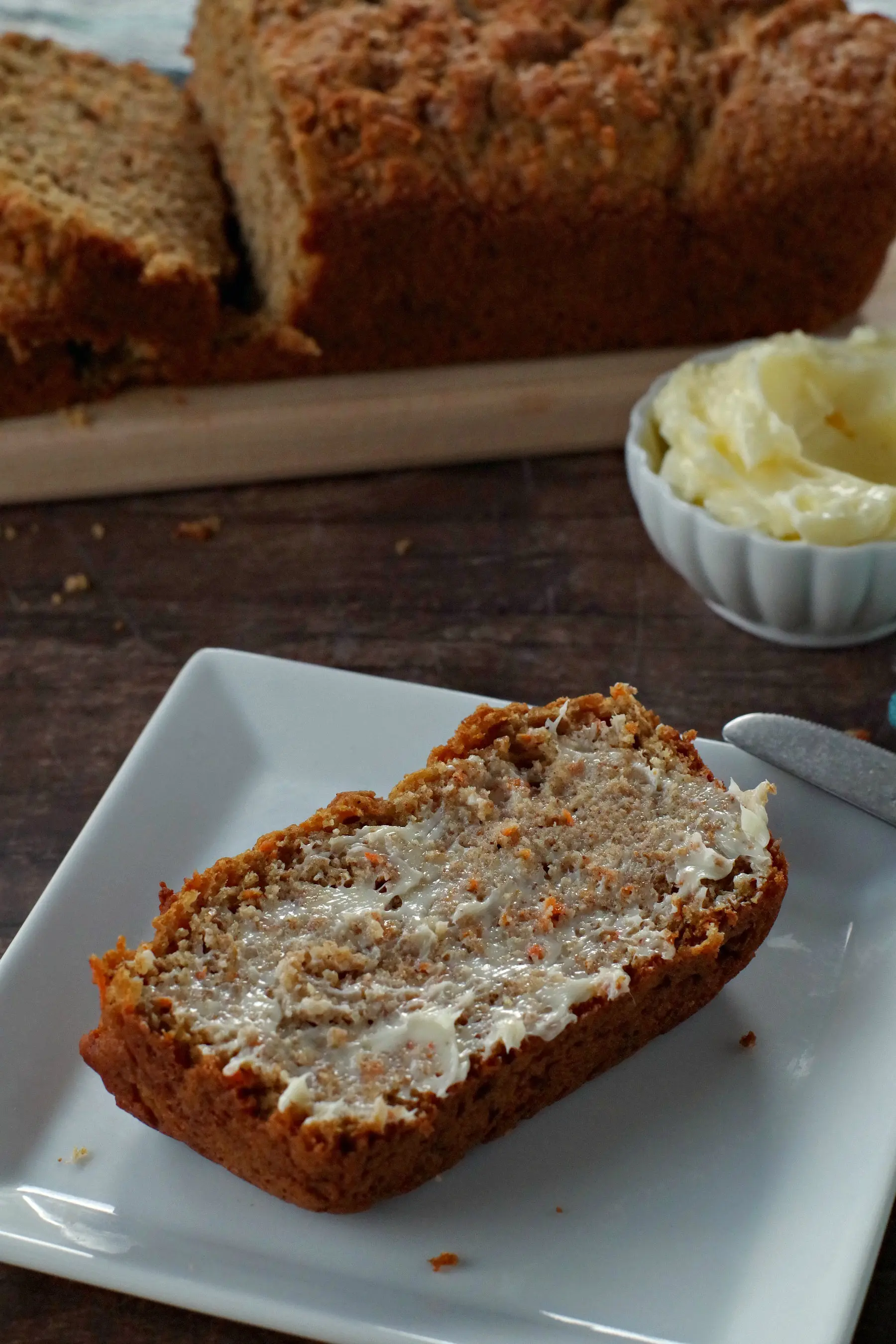 healthy carrot loaf slice with butter on a white plate with white dish of butter in the background. There is also a narrow wooden bread board in the background with the remainder of the carrot loaf on it (partially sliced)