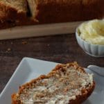 Pin with text at the bottom and top and a photo of healthy carrot loaf slice with butter on a white plate with white dish of butter in the background. There is also a narrow wooden bread board in the background with the remainder of the carrot loaf on it (partially sliced)