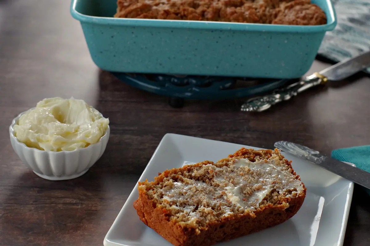 healthy carrot loaf slice with butter on a white plate with white dish of butter in the background. There is also a blue loaf pan in the background with the remainder of carrot loaf in it, as well as a silver knife beside the loaf pan.
