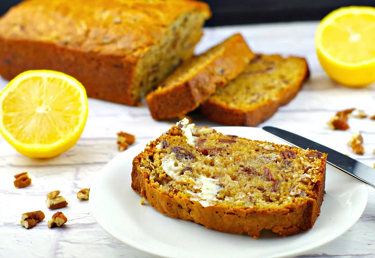 a slice of date loaf on a white plate with whole date loaf in the background (partially sliced) and sliced lemons on the side