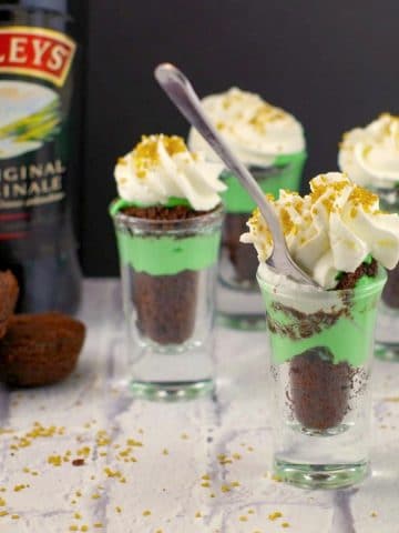 Irish cream dessert shots with a bottle of irish cream in the background, on a white faux surface