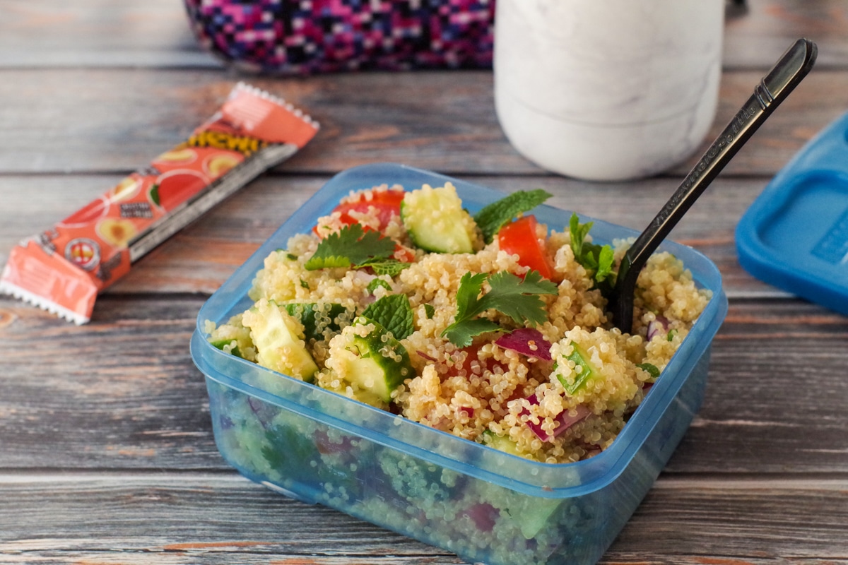 quinoa salad in a blue lunch container with plastic fork and lunch bag, water bottle and granola bar in the background