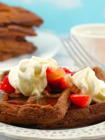 chocolate waffles with strawberries and whipped cream on a white plate white with a stack of waffles in background