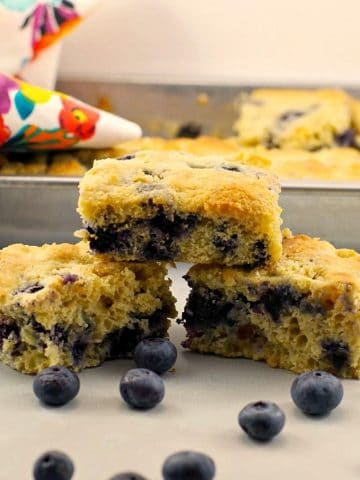 3 pieces of blueberry cornbread stacked on a counter with a pan of blueberry cornbread in background