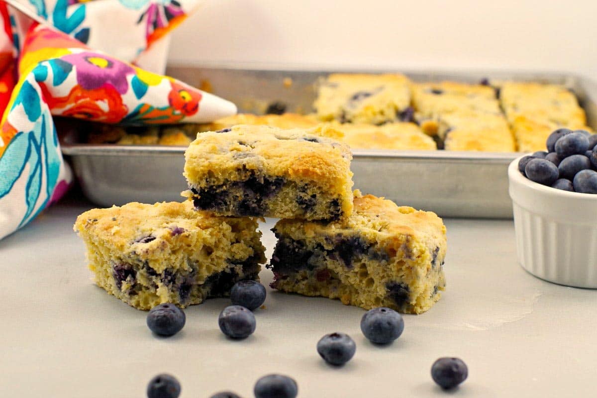 3 pieces of blueberry cornbread stacked on a counter with a pan of blueberry cornbread in background