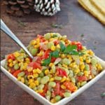 Pinterest Pin with text at top and bottom and photo of corn succotash in a white square bowl (with spoon in it) on brown surface, and pine cones in the background