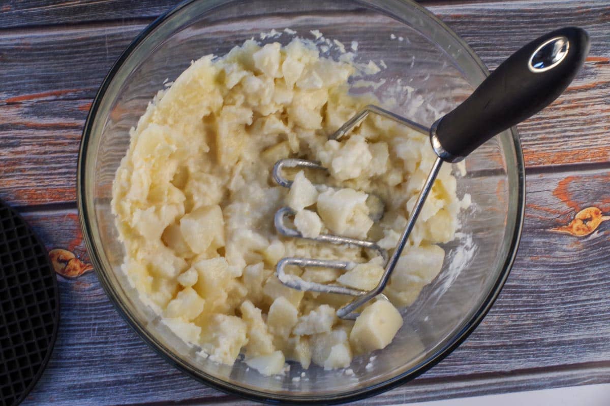 Potatoes being mashed with other ingredients in a large glass bowl.