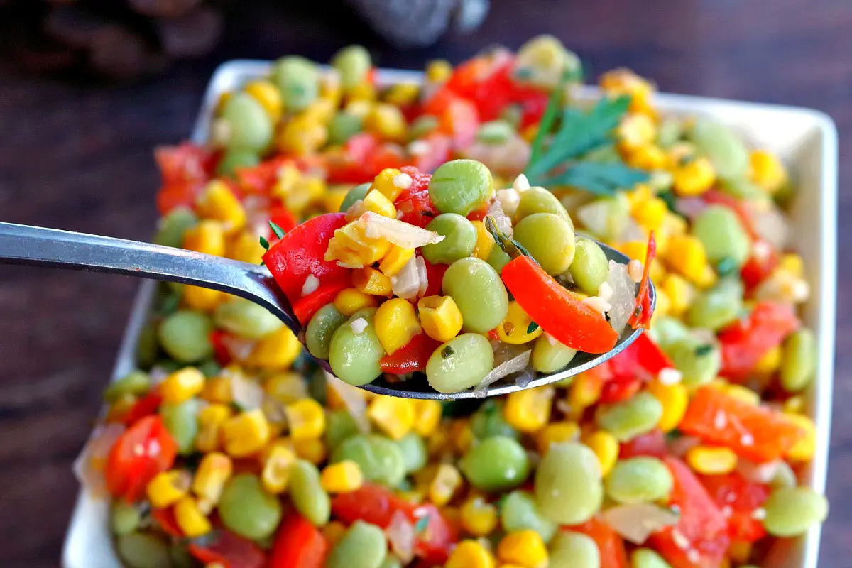 corn succotash being held up on a silver spoon with a dish of succotash below