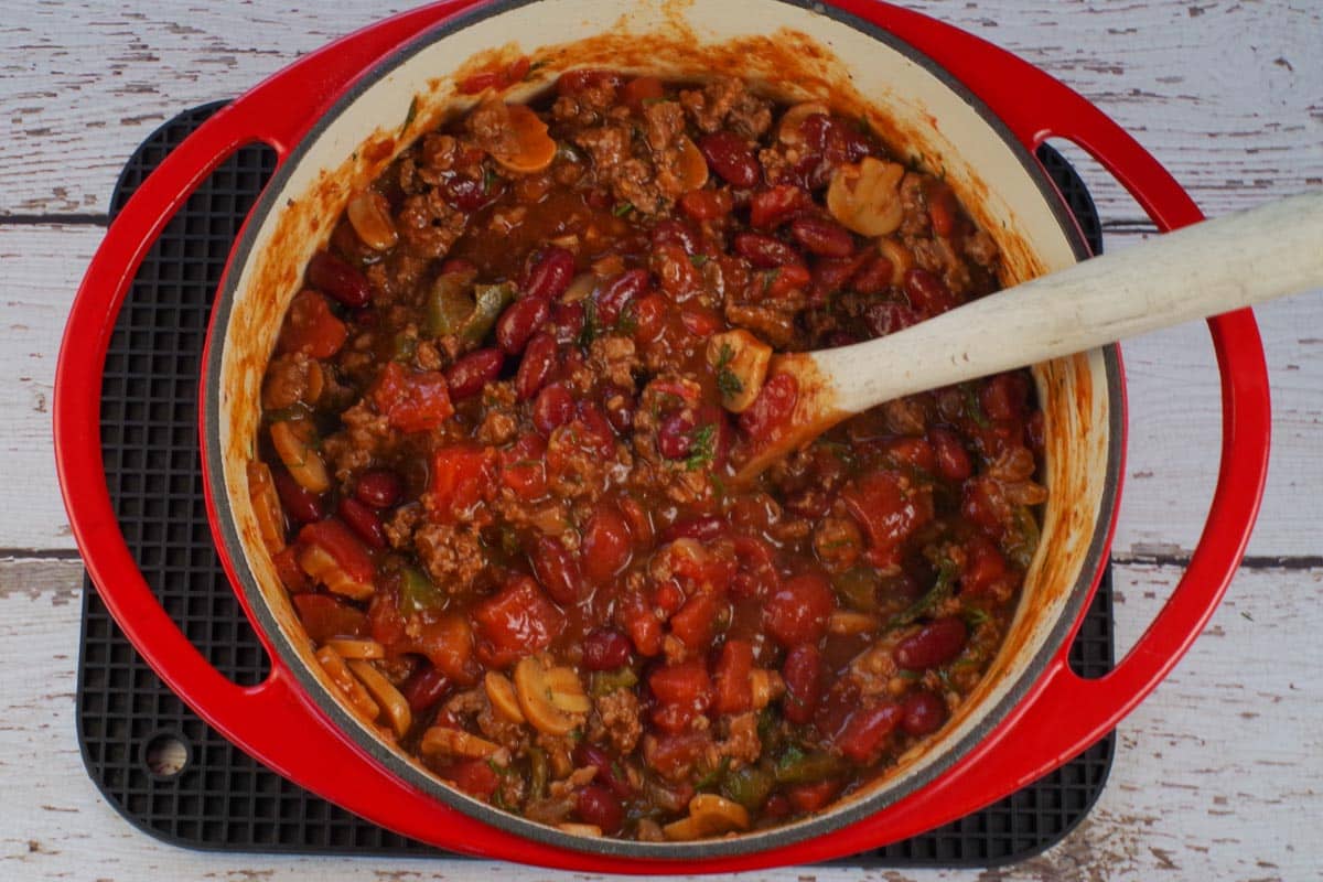 chili in orange dutch oven with wooden spoon with green peppers and onions cooked through