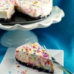 a slice of birthday cake cheesecake on a white plate with a fork on the side and the remainder of the cake on a white stand in the background