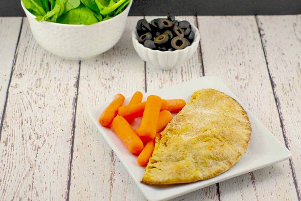 a healthy pizza pocket on a white plate with baby carrots and a dish of olives and spinach in the background