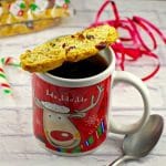 Christmas coffee mug with piece of biscotti on top, spoon on the side and ribbon in the background