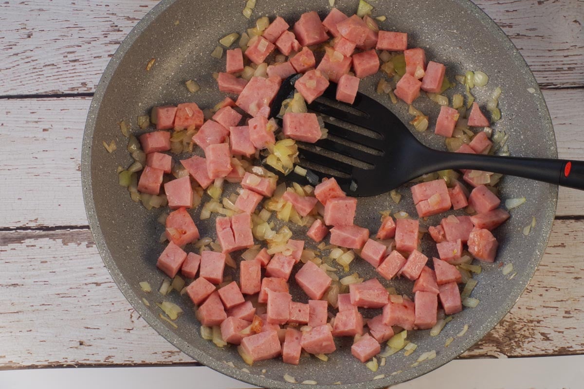 Cubed ham and onions in skillet with black spatula.