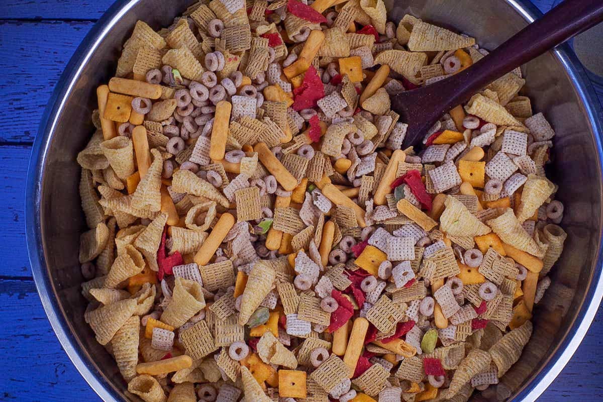 Cheerios, Life cereal, Chex cereal, pretzels (missing in photo), Bugles, pistachios, cheese nips, cheese bits, and red tortilla chips mixed together in large stainless steel bowl.