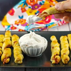 breakfast pigs in a blanket on black tray with dip in the middle and one being dipped into dip