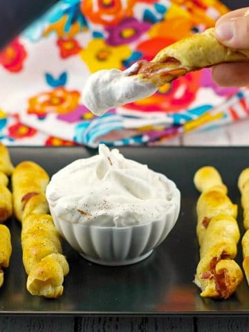 breakfast pigs in a blanket on black tray with dip in the middle and one being dipped into dip