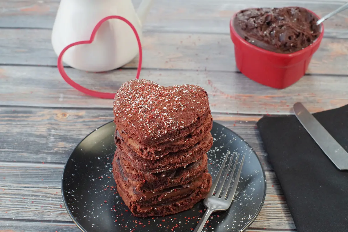 stack of 6 healthy red velvet pancakes on a black plate with a fork on the side, heart-shaped dish with chocolate cream cheese butter and white syrup container with heart shaped cookie cutter leaning against it, in the background