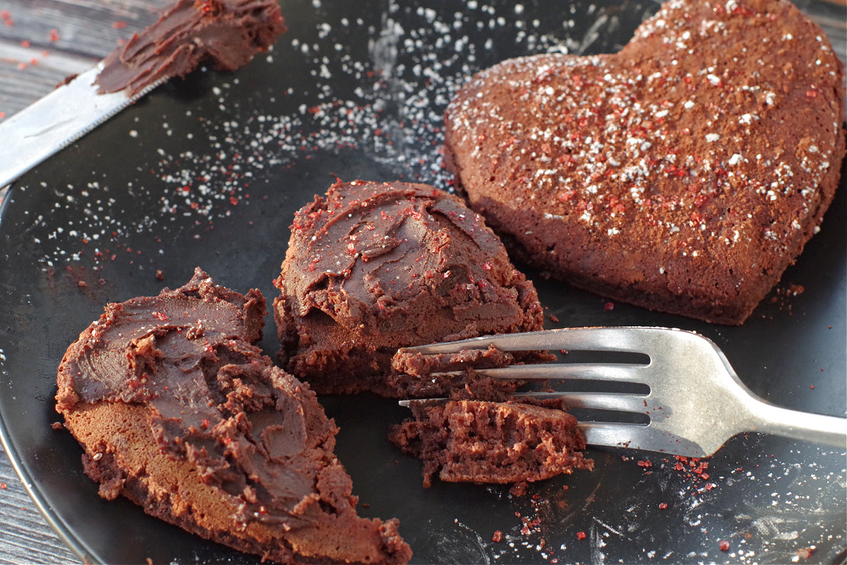 2 heart shaped red velvet pancakes on a black plate, one with chocolate cream cheese butter being cut into and the other with red sparkling sugar and powdered sugar