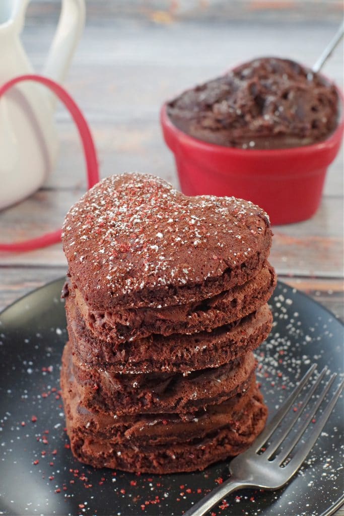 stack of 6 healthy red velvet pancakes on a black plate with a fork on the side, heart-shaped dish with chocolate cream cheese butter, in the background
