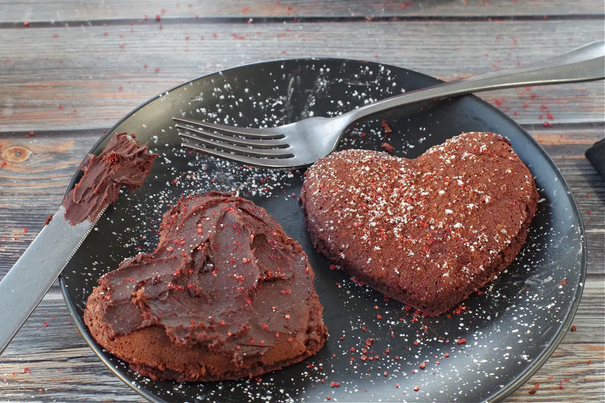 2 heart shaped red velvet pancakes on a black plate, one with chocolate cream cheese butter and the other with red sparkling sugar and powdered sugar