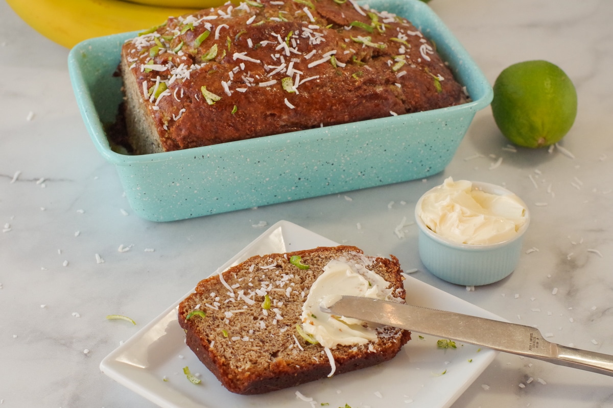 banana bread in blue loaf pan with slice of banana bread on a white plate with butter