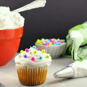 Frosted cupcake with pearl sprinkles, spatula and decorating tip in foreground with orange bowl of frosting and small bowl pearl sprinkles in background