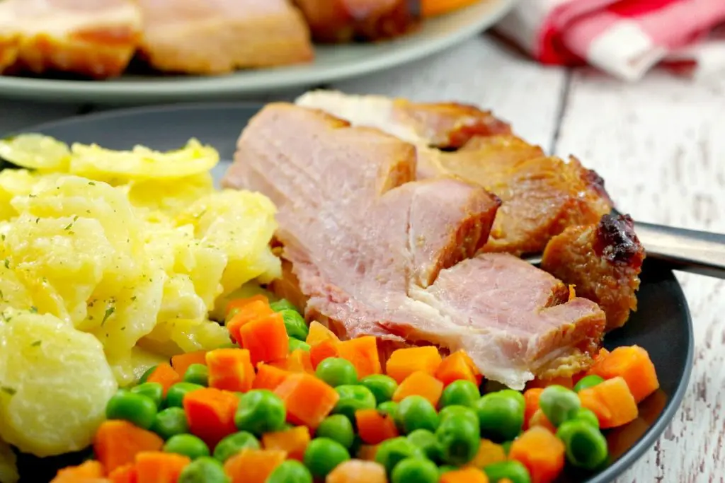 Sliced picnic ham on a plate with scalloped potatoes and veggies with whole ham in background