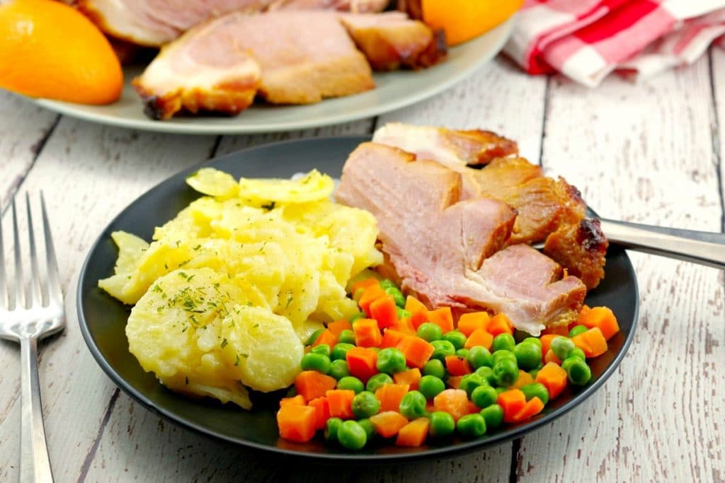 Sliced picnic ham on a plate with scalloped potatoes and veggies with whole ham in background