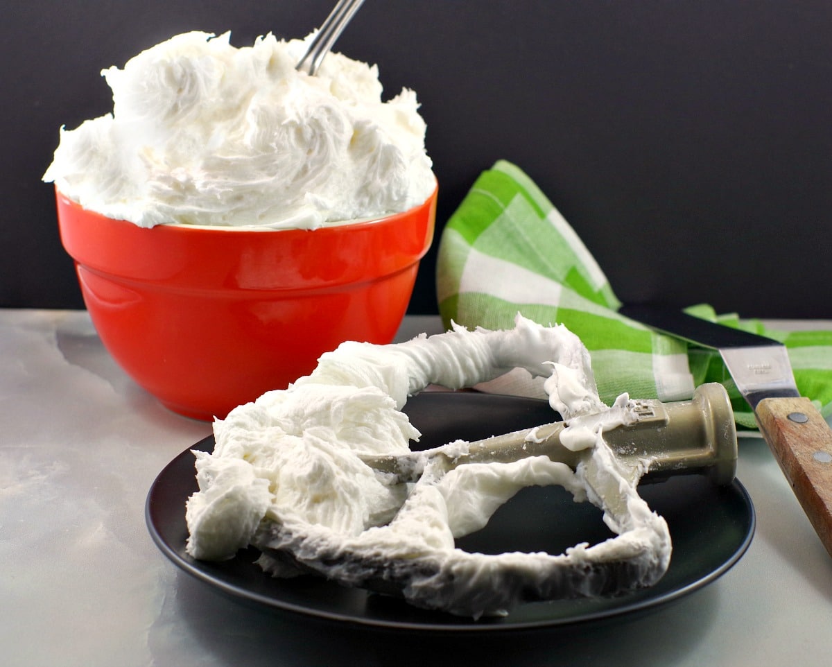 Stand mixer paddle attachment with icing on it in the foreground (on plate) and orange bowl of whipped frosting in background