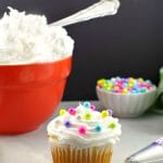 Frosted cupcake with pearl sprinkles, spatula and decorating tip in foreground with orange bowl of frosting and small bowl pearl sprinkles in background