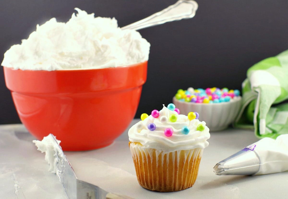 Frosted cupcake with pearl sprinkles, spatula and decorating tip in foreground with orange bowl of frosting and small bowl pearl sprinkles in background