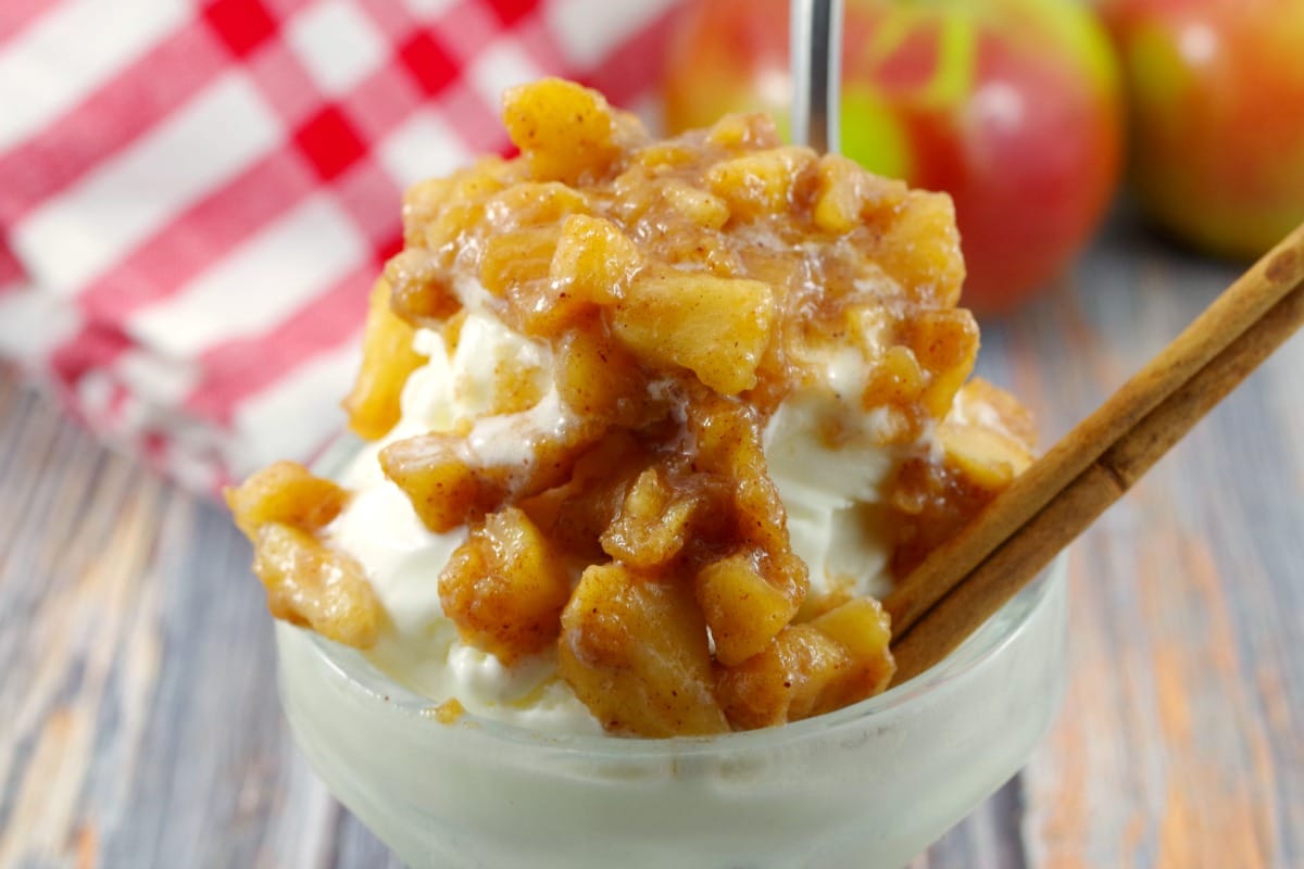 apple cinnamon topping on frozen yogurt in ice cream dish with spoon and cinnamon stick, apple and checked cloth in background