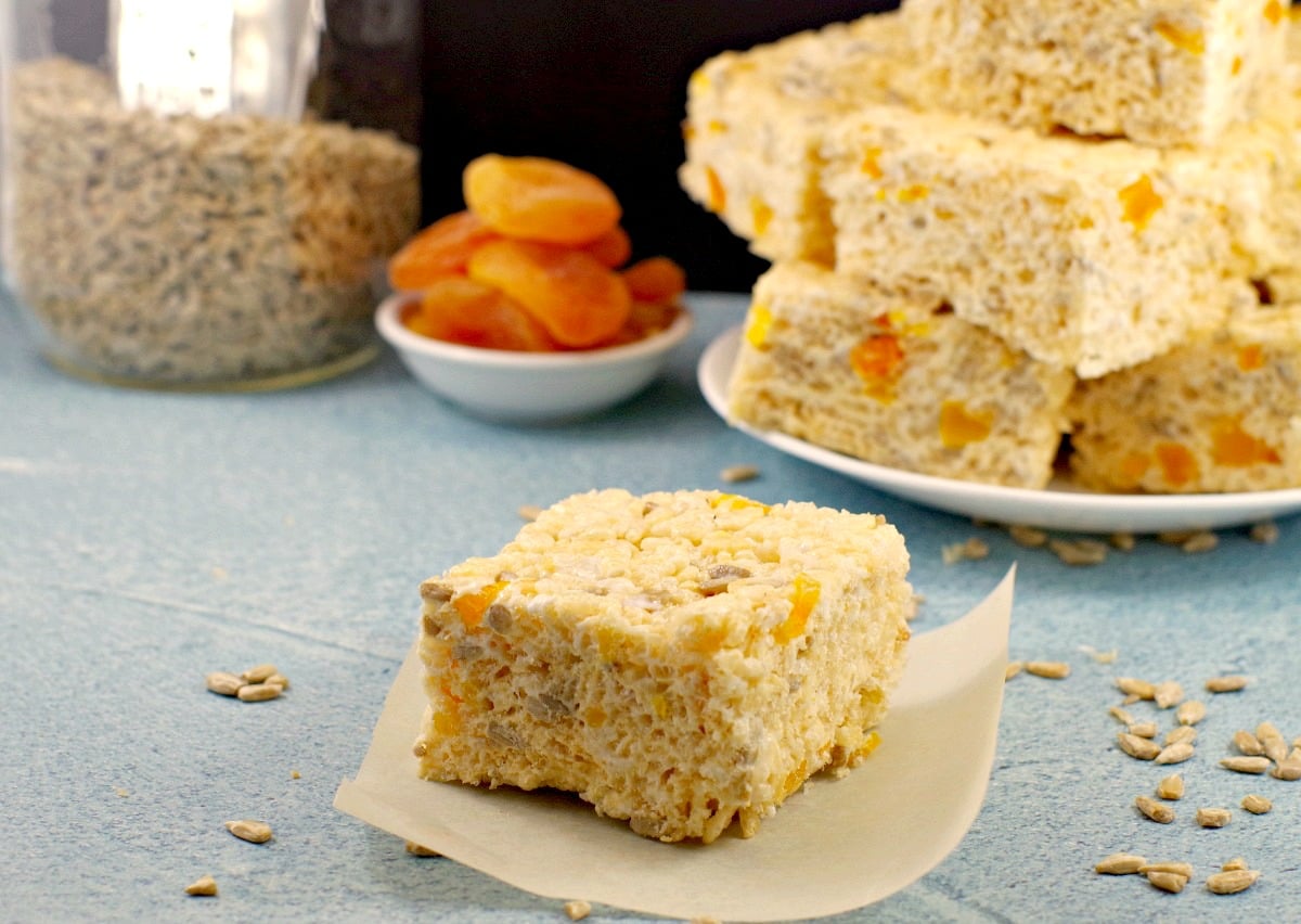 Rice Krispie treat on parchment paper with plate of treats, bowl of apricots and jar of sunflower seeds in the background