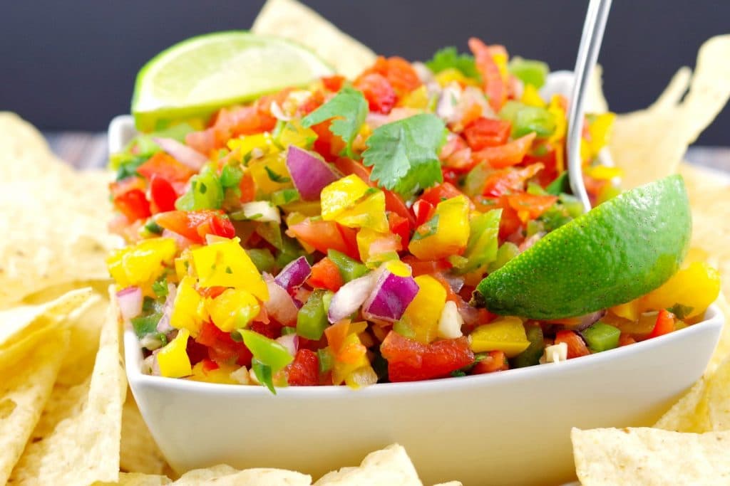 Pico de Gallo Salsa. garnished with 2 lime wedges, in a white bowl surrounded by nacho chips