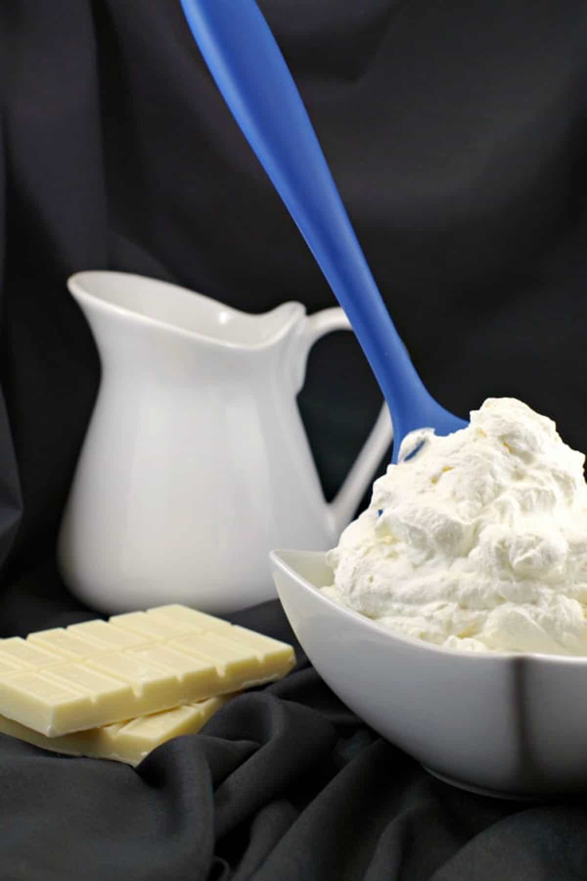 bowl of white chocolate whipped cream frosting with blue spatula handle sticking out and white cream container in background